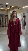 Front view of model wearing burgundy pleated maxi evening gown with cape sleeves and front cutout