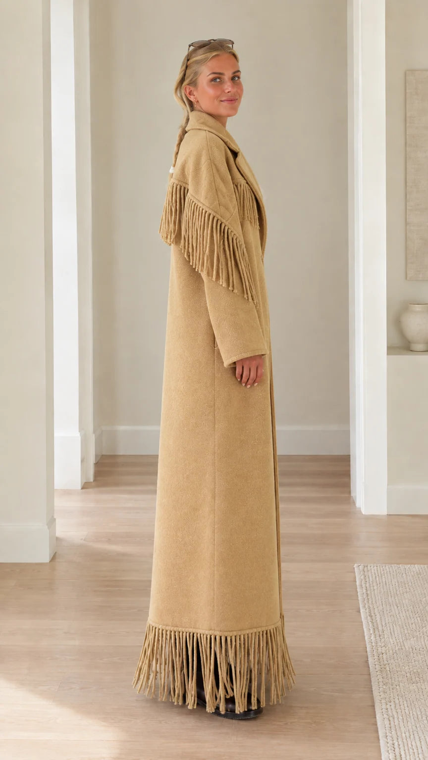 Side profile view of woman wearing long camel overcoat with shoulder and hem fringe